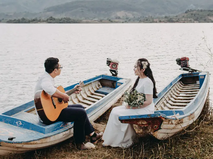 A man playing guitar for a bride, sitting between two wooden boats on a lake